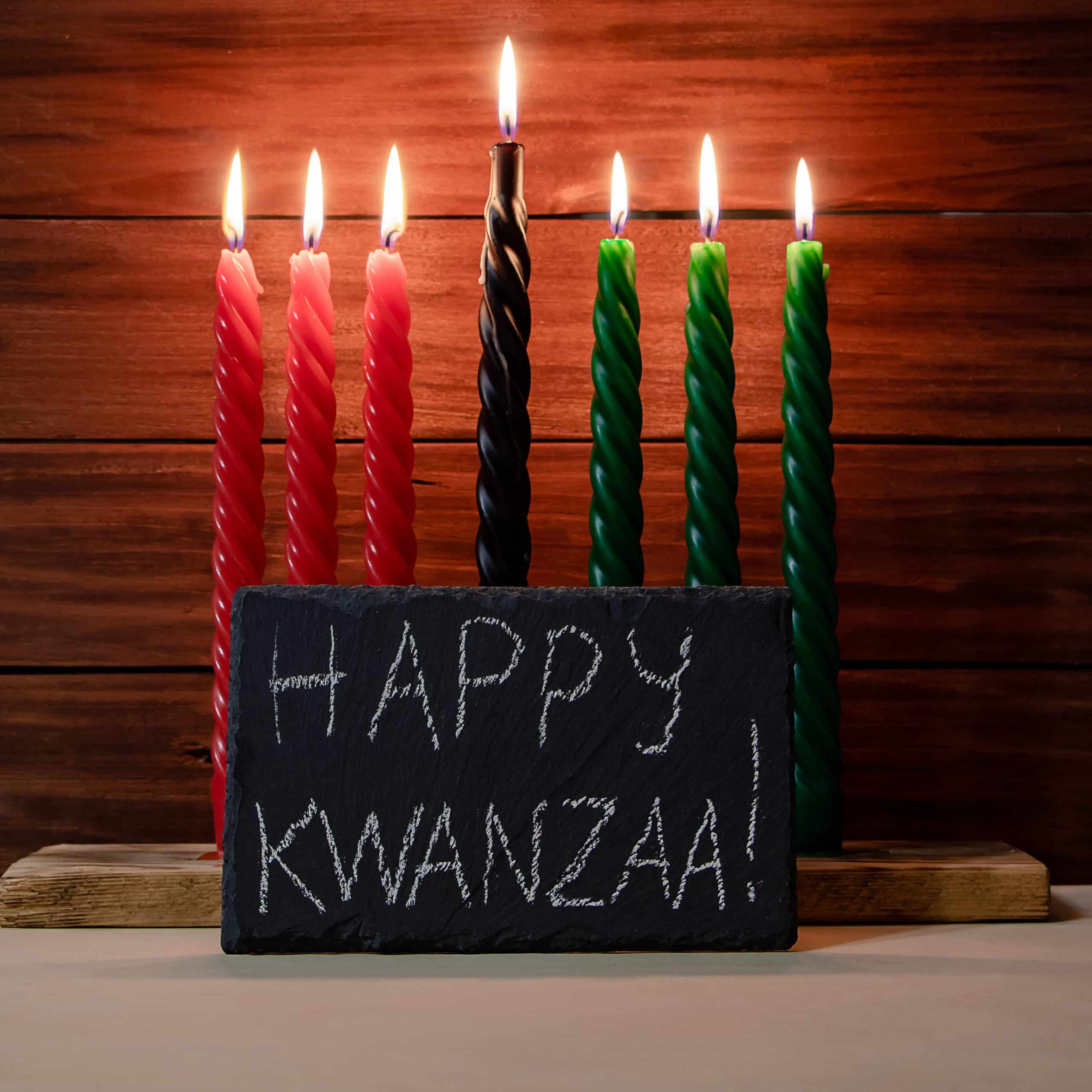 Happy Kwanzaa. African American holiday. Seven candles red, black and green on wooden background. Symbols of African heritage. Congratulatory inscription on chalk board.